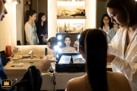 Behind the Scenes: Bride Gets Wedding-Ready on Cape Fahn Island in Samui On an island at Cape Fahn, Samui, a bride sits in her wedding attire, gazing into a mirror surrounded by helpers amidst the glow of light bulbs.